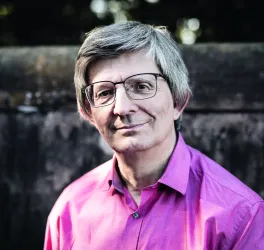 Ein lächelnder Mann mit grauen Haaren trägt eine pinke Hemd und Brille. Im Hintergrund ist eine unscharfe Mauer zu sehen.