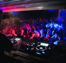 Ein DJ spielt Musik vor einer großen, tanzenden Menschenmenge. Bunte Lichter und ein kreatives Ambiente schaffen eine lebendige Partystimmung.