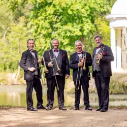 Vier Musiker stehen am Ufer eines Teichs und halten Posaunen in der Hand. Sie tragen elegante Anzüge mit Fliegen und lächeln in die Kamera.