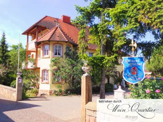 Ein charmantes Gebäude mit einem roten Dach, umgeben von Bäumen und blühenden Pflanzen. Neben dem Eingang steht ein blaues Schild mit einem Wappen.