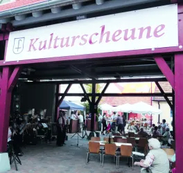 Eine Veranstaltung in der Kulturscheune mit Sitzplätzen und einer Band. Menschen stehen und sitzen zusammen unter einem Vordach.