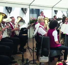 Eine Blaskapelle spielt unter einem Zelt. Die Musiker tragen rote Westen und konzentrieren sich auf ihre Instrumente.