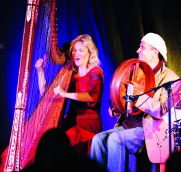 Zwei Musiker auf der Bühne, eine Frau spielt Harfe und ein Mann ein Handtrommeln. Die Atmosphäre ist lebhaft und musikalisch.