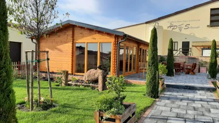 Ein gemütliches Holzhaus mit großen Fenstern steht in einem gepflegten Garten. Umgeben von Bäumen und straff geschnittenen Hecken wirkt die Umgebung einladend und ruhig.
