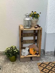 Eine Holzpalette dient als kleines Regal, dekoriert mit einer Laterne, einer Blumentopf und verschiedenen Gegenständen. Daneben steht ein Topf mit blühenden Pflanzen.