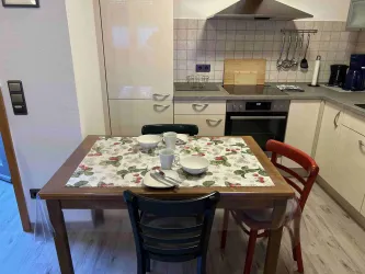 Eine helle Küche mit einem Tisch, der mit Geschirr und einer Tischdecke dekoriert ist. Es gibt drei Stühle in verschiedenen Farben und moderne Küchengeräte im Hintergrund.