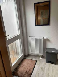 Ein heller Eingangsbereich mit einer weißen Tür und einem Spiegel. Ein Heizkörper und ein Mülleimer sind ebenfalls sichtbar.