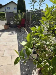 Ein sonniger Außenbereich mit Terrassenfliesen und Bepflanzung. Im Hintergrund stehen ein paar Töpfe und ein bequemer Stuhl.