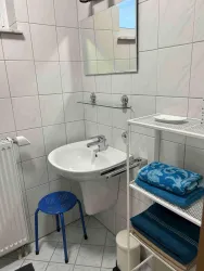 Ein kleines Badezimmer mit weißen Fliesen und einem Waschbecken. Es gibt einen blauen Hocker und Regale mit Handtüchern.