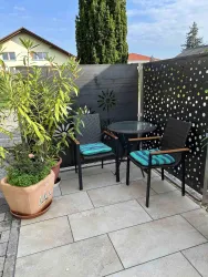 Ein gemütlicher Außenbereich mit einem kleinen Tisch und zwei Stühlen. Im Hintergrund steht ein Pflanze und eine dekorative Trennwand.