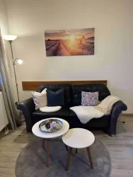 Ein gemütliches Wohnzimmer mit einem schwarzen Sofa und einem kleinen Tisch. An der Wand hängt ein Bild mit einer Sonnenuntergangslandschaft.