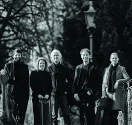 Eine Gruppe von fünf Musikern steht auf einer Brücke umgeben von Bäumen. Sie halten Instrumente und schauen in die Kamera.