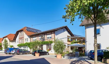 Ein malerisches Dorf mit modernen und traditionellen Häusern. Die Straße ist mit Bäumen gesäumt, und es stehen einige Autos parat.