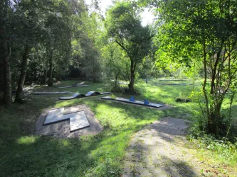 Ein grüner Park mit schattenspendenden Bäumen und einem speziellen Bewegungsbereich. Es gibt verschiedene Bewegungsstationen auf dem Rasen.