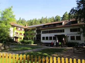 Ein zweigeschossiges Gebäude mit bunten Fassaden umgeben von Bäumen. Vor dem Gebäude sind Bänke und ein gepflegter Rasen zu sehen.