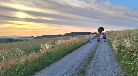 Eine Gruppe von Menschen geht einen Feldweg entlang. Der Sonnenuntergang färbt den Himmel in warmen Farben.