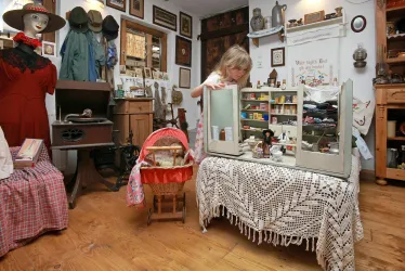 Ein nostalgischer Raum mit einem Kind, das an einem Puppenhaus spielt. Im Hintergrund sind alte Möbel und verschiedene Ausstellungsstücke zu sehen.