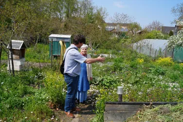 Zwei Personen stehen in einem blühenden Garten und unterhalten sich. Im Hintergrund sind andere Gartenstrukturen und viele Pflanzen sichtbar.