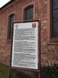 Ein Informationsschild steht an einer historischen Gebäudeaußenwand. Es enthält Texte und Wappen, die über die Geschichte des Ortes informieren.