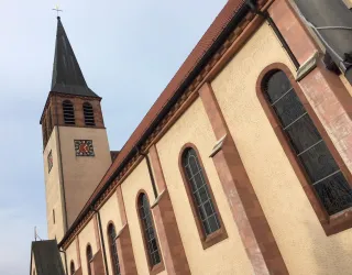 Eine Kirche mit einem hohen Turm und einem Zifferblatt. Die Fassade ist aus hellem Stein und die Fenster sind teilweise verziert.
