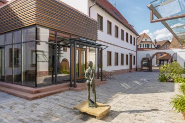 Ein moderner Anbau mit Glasfront und Holzverkleidung an einem historischen Gebäude. In der Mitte steht eine Statue auf einem Holzsockel, umgeben von einem gepflasterten Innenhof.