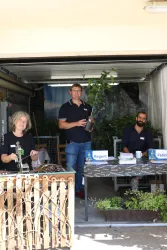 Drei Personen arbeiten in einer Garage. Ein Mann hält eine Pflanze, während zwei andere an einem Tisch sitzen.