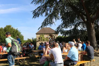 Eine lebendige Gartenparty mit vielen Menschen, die an Tischen sitzen und sich unterhalten. Im Hintergrund ist ein großes Gebäude und Bäume zu sehen.