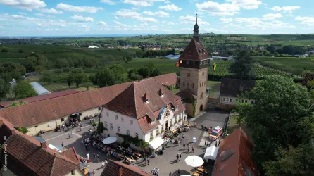 Ein historisches Gebäude mit einem hohen Turm und roten Dächern. Der Innenhof ist belebt und von einer schönen Landschaft umgeben.