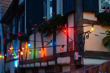 Ein Haus mit traditioneller Fachwerkarchitektur, geschmückt mit bunten Lichtern. Blumen im Fenster und eine einladende Atmosphäre schaffen eine gemütliche Stimmung.