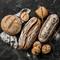 Eine Auswahl an frisch gebackenem Brot auf einem dunklen Untergrund. Dazu gehören runde und längliche Brote sowie Brötchen, umgeben von einem grauen Tuch.