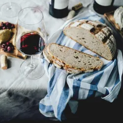 Ein Glas Rotwein steht neben frisch gebackenem Brot auf einem gedeckten Tisch. Dazu gibt es verschiedene Häppchen und eine gestreifte Tischdecke.