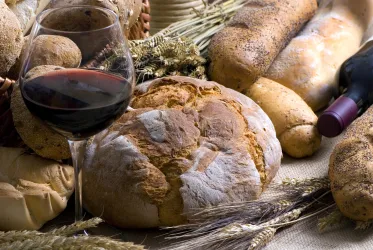 Ein rustikaler Tisch mit verschiedenen Brotsorten und einem Glas Rotwein. Dazu liegen Ähren und eine Flasche Wein.
