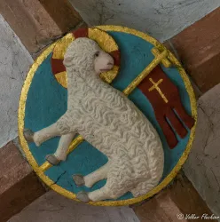 Ein weißes Lamm mit einer Fahne steht auf einem blauen Hintergrund. Es ist von einem goldenen Rand umgeben und hat einen Heiligenschein.