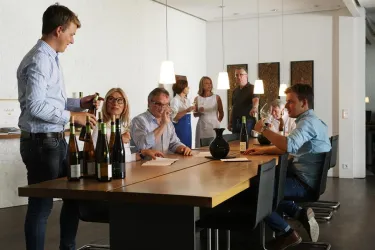 Eine Gruppe von Menschen sitzt an einem langen Tisch und genießt Wein. Einige Personen sprechen miteinander, während andere den Wein betrachten.