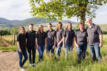 Eine Gruppe von acht Personen steht auf einem Feld mit Weinreben. Im Hintergrund sind Hügel und ein klarer Himmel sichtbar.