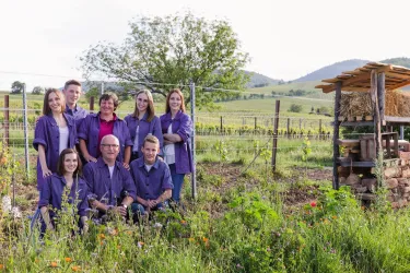Eine Gruppe von Menschen steht in einem Garten mit Blumen und Pflanzen. Im Hintergrund sind sanfte Hügel und eine Scheune zu sehen.