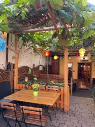 Ein gemütlicher Außenbereich mit einem Holztisch und Stühlen. Über dem Tisch hängen Lichter und Weinreben, die für eine einladende Atmosphäre sorgen.