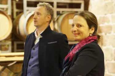Ein Mann und eine Frau stehen in einem Weinkeller zwischen Eichenfässern. Die Frau lächelt freundlich, während der Mann nachdenklich aussieht.