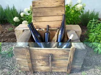 Eine Holzkiste mit mehreren Flaschen steht im Garten. Im Hintergrund sind grüne Pflanzen und Blüten zu sehen.