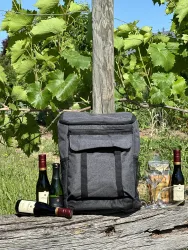 Ein schwarzer Rucksack steht auf einem Holzstamm, umgeben von Weinflaschen und Gläsern. Im Hintergrund sind grüne Weinreben und ein blauer Himmel zu sehen.