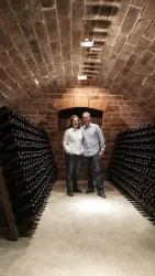 Ein Paar steht in einem Weinkeller, umgeben von vielen Weinflaschen. Die Wände sind aus rotem Stein und die Beleuchtung ist sanft.