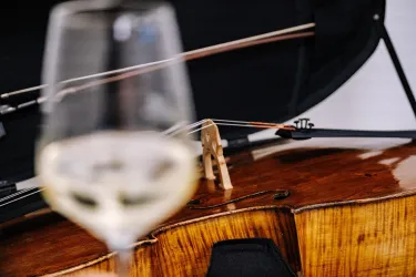 Ein elegantes Glas Weißwein steht im Vordergrund, während im Hintergrund ein Cello zu sehen ist. Die detailreiche Holzmaserung des Cellos ist gut erkennbar.