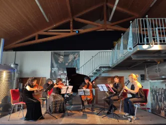 Ein Ensemble aus sechs Musikern spielt in einem modernen Raum. Im Hintergrund sind große Poster und ein Klavier zu sehen.