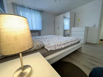 Ein gemütliches Schlafzimmer mit einem Bett, einem Nachttisch und einer Lampe. Die Wände sind hell und es gibt einen großen Schrank sowie ein Fenster mit Vorhängen.
