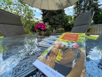 Ein Tisch im Freien mit einem Buch und einer Vase mit blühenden Pflanzen. Im Hintergrund sind Bäume und ein Sonnenschirm zu sehen.