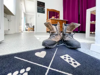 Ein Paar schmutzige Arbeitsstiefel stehen auf einem Fußmatte mit einem einfachen Muster. Im Hintergrund sind ein gemütlicher Raum und ein Spiegel zu sehen.