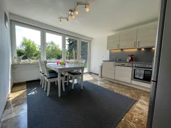 Eine helle, moderne Küche mit einem Essbereich. Große Fenster bieten einen Blick auf den Garten.