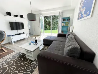 Ein modernes Wohnzimmer mit einem grauen Sofa, einem Fernseher und großen Fenstern. Der Raum hat eine helle Atmosphäre und einen Blick auf den Garten.