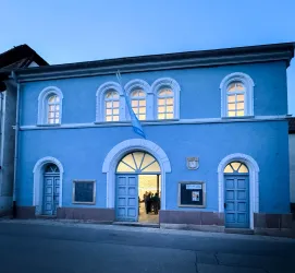 Ein blaues Gebäude mit großen Fenstern und zwei Eingangstüren. Die Fassade ist einfach und modern, mit einem leichtem Lichtschein im Inneren.