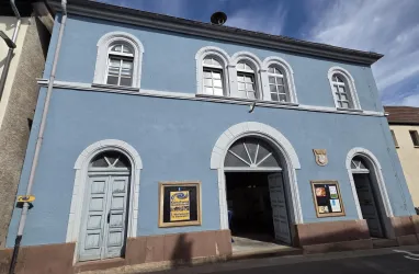 Ein blaues Gebäude mit mehreren Fenstern und Türen. Die Fassade hat einen klassischen Stil und bietet Platz für Informationen an der Wand.